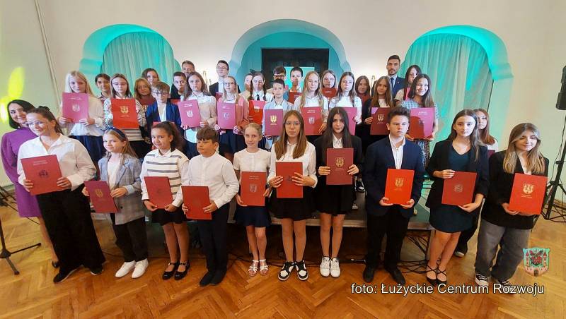 Na pierwszym planie szykownie ubrane dzieci, które w dłoniach trzymają bordowe kartki papieru ze złotymi zdobieniami. W tle białe ściany z niebieskim podświetleniem.